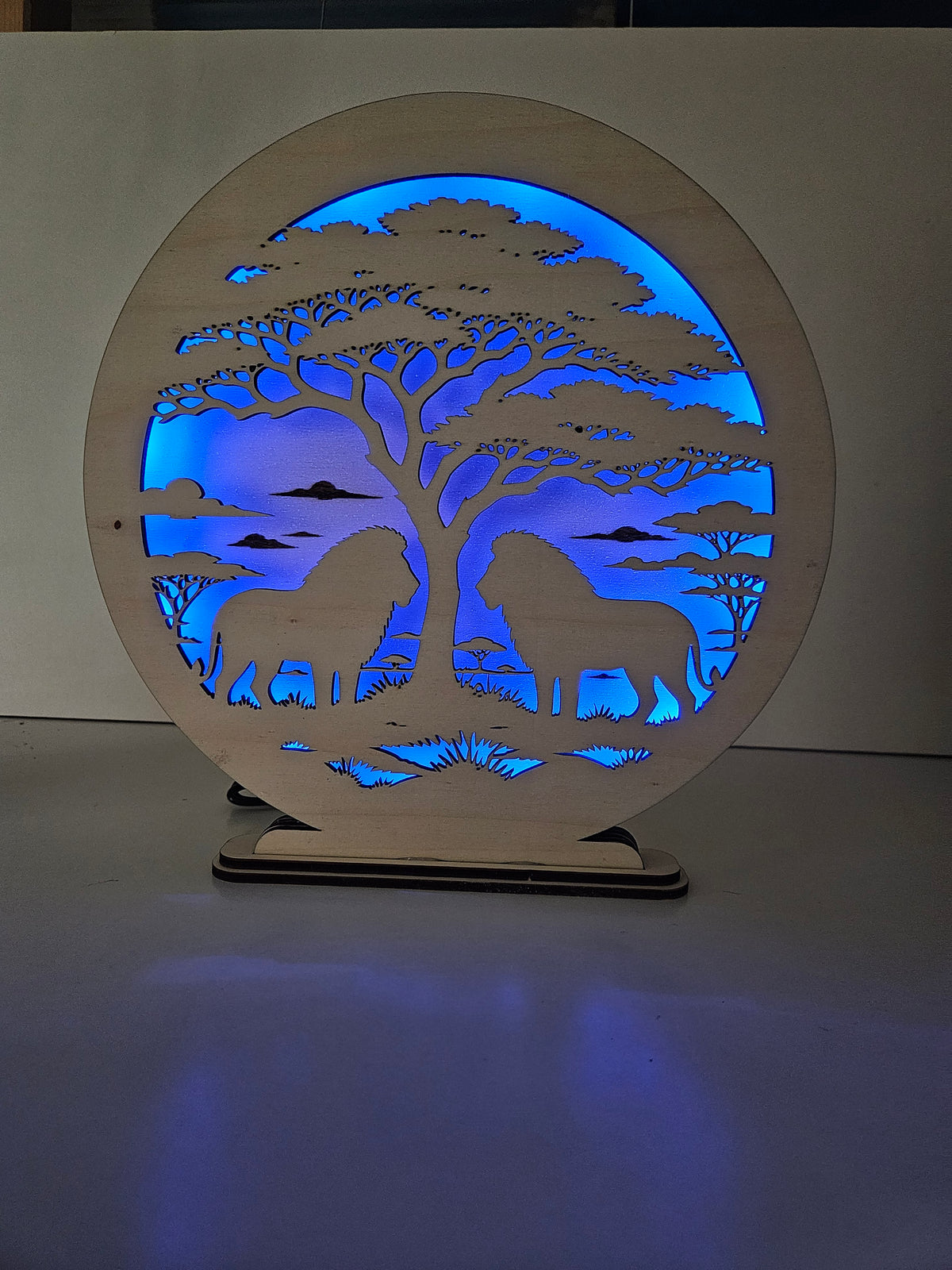 Runde Holzlampe mit Savannenmotiv, zwei Löwen unter einer Akazie, blau beleuchtet von RGB-LEDs